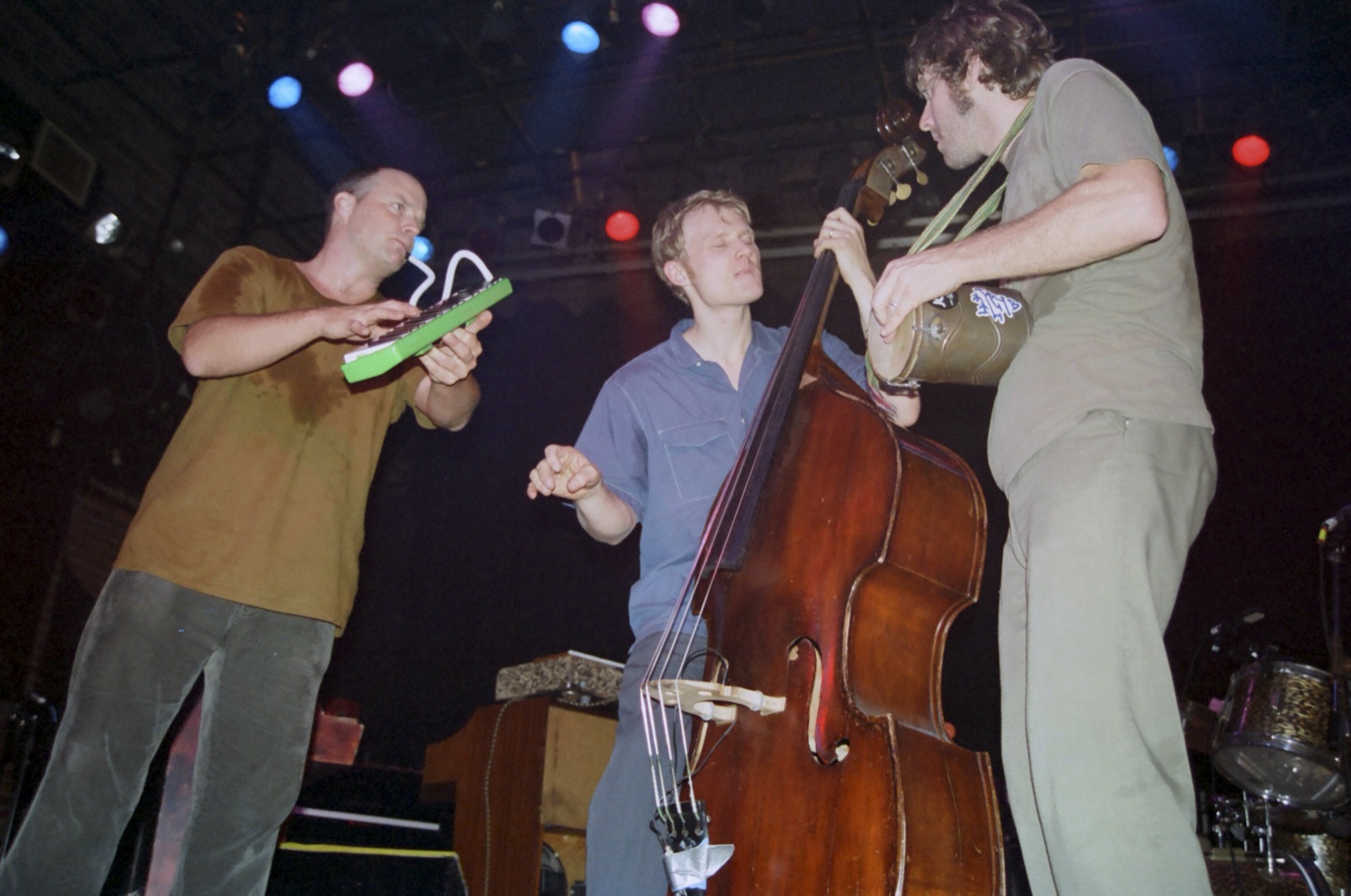 Three musicians crowd together at the front of the stage with a melodica, an upright bass and a hand drum under colored spotlights.